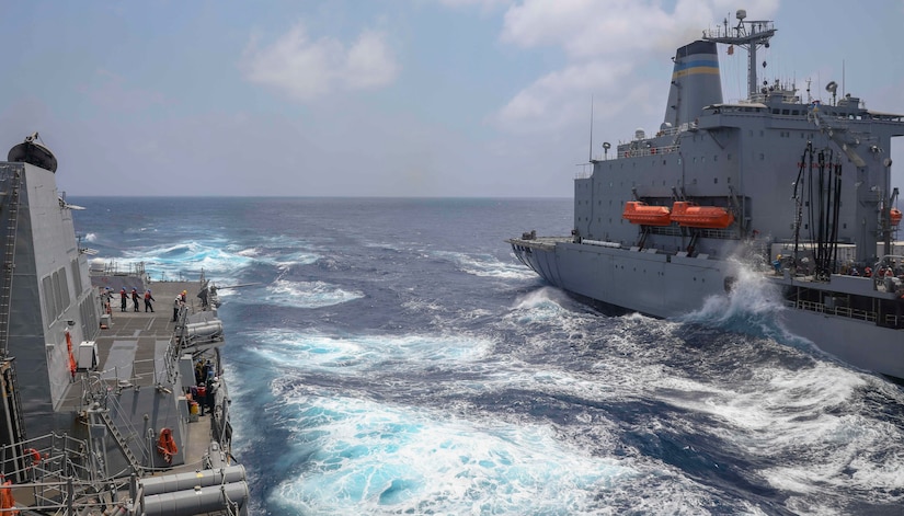 Two ships are alongside each other at sea; a cable is extended from one to the other and waves crash below.