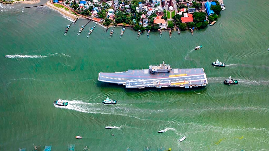 An aerial shot shows a large aircraft carrier being escorted by smaller vessels through a narrow pass.