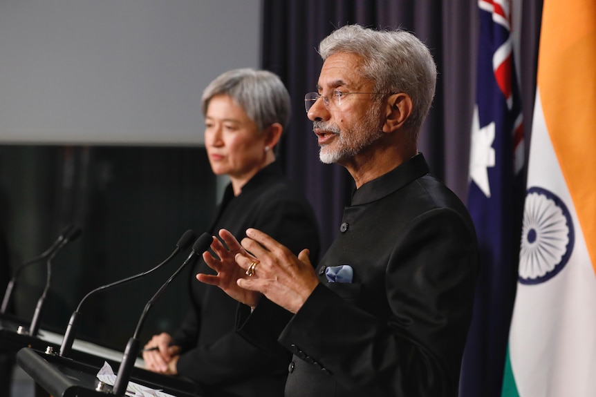 A man and woman stand on a stage giving a press conference with the Australian and Indian flags behind them. 